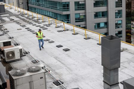 Roof maintenance engineer doing a routine inspection on the rooftop of a building and taking notes on his tablet
