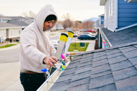 A hispanic man standing on a ladder, hanging Christmas lights on the outside of his home.