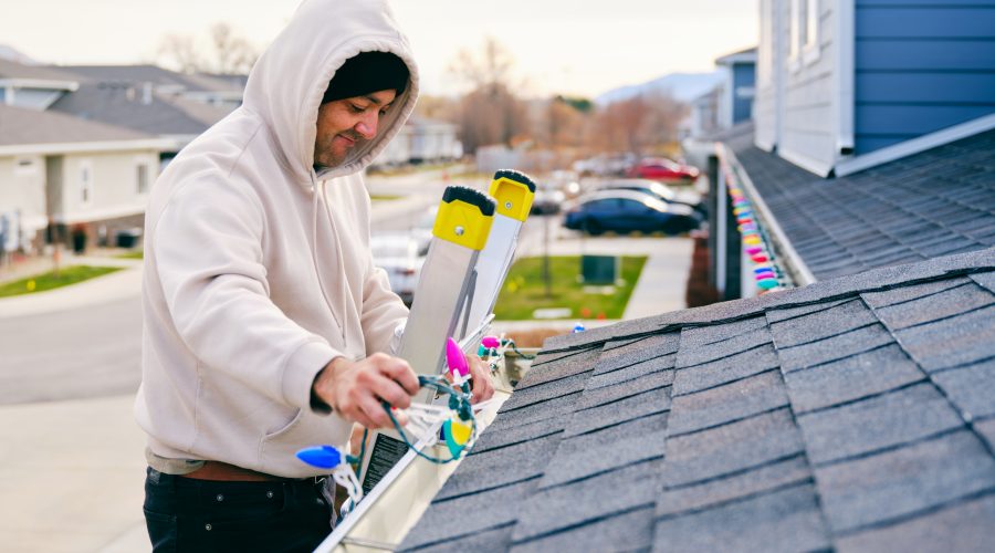 A hispanic man standing on a ladder, hanging Christmas lights on the outside of his home.